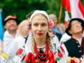 Młoda kobieta w tradycyjnym stroju ludowym z kwiatami we włosach i koralami. Obok ludzie w strojach, biało-czerwone flagi. Świętujemy 16 sierpnia!