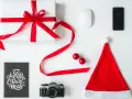 Świąteczne prezenty dla informatyka: białe pudełko z czerwoną wstążką, myszka, iPhone, aparat, kartka "Merry Christmas" i czapka Mikołaja.