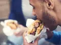 Mężczyzna z zarostem zajada soczystego burgera, świętując dzień burgera. W tle widać drugą osobę z podobnym przysmakiem.