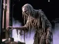 Stary, obdarty bieda demon w szarych szatach, z ćmą na ramieniu, wyciąga rękę do okna.