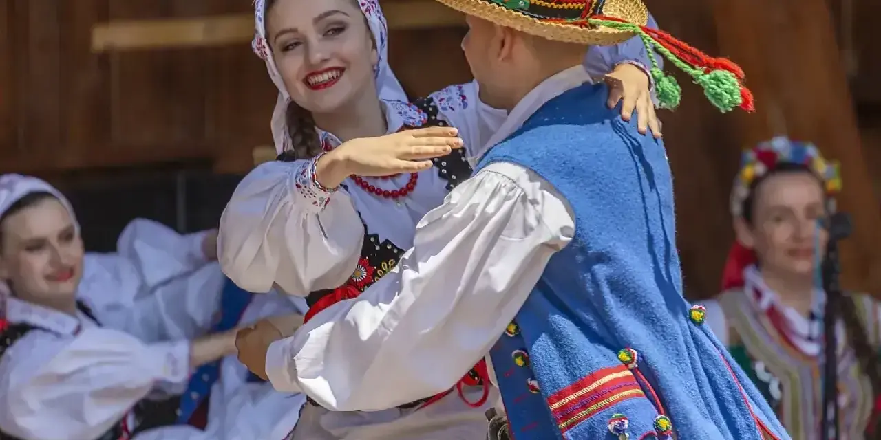 Młoda para w strojach ludowych tańczy z uśmiechem. Chłopak w słomkowym kapeluszu, dziewczyna w kwiecistej chustce. Licho co to za piękny widok!