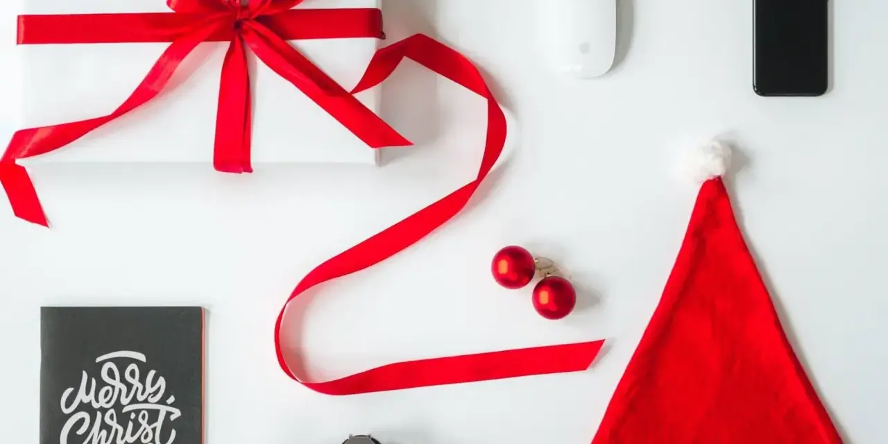 Świąteczne prezenty dla informatyka: białe pudełko z czerwoną wstążką, myszka, iPhone, aparat, kartka "Merry Christmas" i czapka Mikołaja.