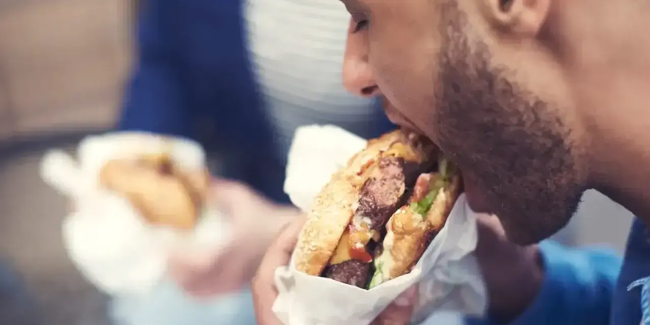 Mężczyzna z zarostem zajada soczystego burgera, świętując dzień burgera. W tle widać drugą osobę z podobnym przysmakiem.