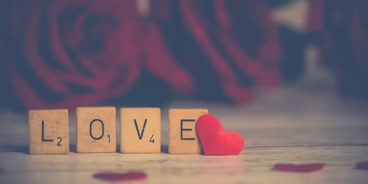 Drewniane klocki z napisem "LOVE" i czerwone serce na tle róż. Od kiedy obchodzimy walentynki w Polsce?