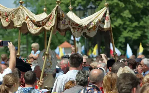Procesja 19 czerwca. Ludzie obserwują uroczystość pod ozdobnym baldachimem, niektórzy filmują telefonami.