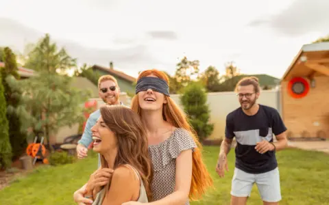 Radosna ciuciubabka zabawa w ogrodzie. Dziewczyna z zawiązanymi oczami śmieje się, podczas gdy jej przyjaciele ją prowadzą.