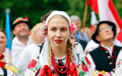 Młoda kobieta w tradycyjnym stroju ludowym z kwiatami we włosach i koralami. Obok ludzie w strojach, biało-czerwone flagi. Świętujemy 16 sierpnia!