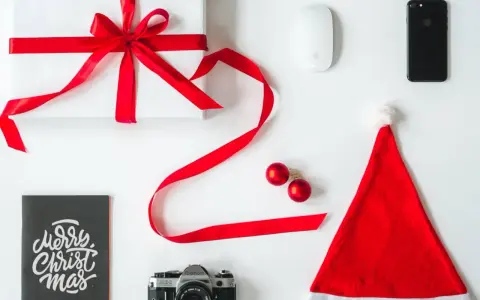 Świąteczne prezenty dla informatyka: białe pudełko z czerwoną wstążką, myszka, iPhone, aparat, kartka "Merry Christmas" i czapka Mikołaja.