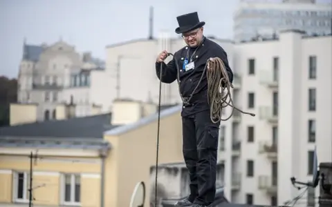 Kominiarz w cylindrze i czarnym stroju przygotowuje się do pracy, trzymając linę. To jego dzień, dzień kominiarza.