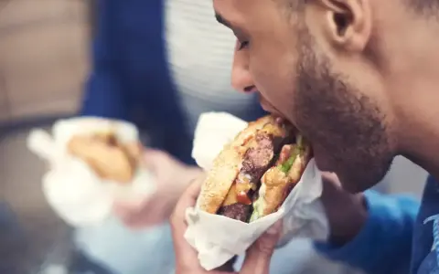 Mężczyzna z zarostem zajada soczystego burgera, świętując dzień burgera. W tle widać drugą osobę z podobnym przysmakiem.