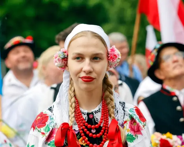 Młoda kobieta w tradycyjnym stroju ludowym z kwiatami we włosach i koralami. Obok ludzie w strojach, biało-czerwone flagi. Świętujemy 16 sierpnia!