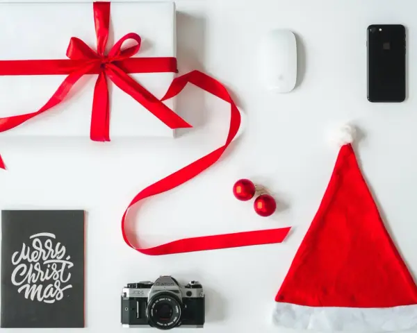 Świąteczne prezenty dla informatyka: białe pudełko z czerwoną wstążką, myszka, iPhone, aparat, kartka "Merry Christmas" i czapka Mikołaja.