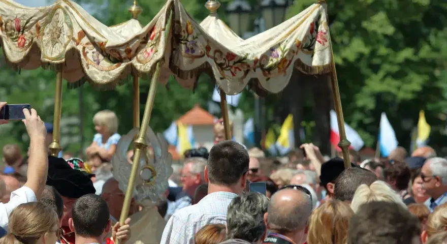 Procesja 19 czerwca. Ludzie obserwują uroczystość pod ozdobnym baldachimem, niektórzy filmują telefonami.