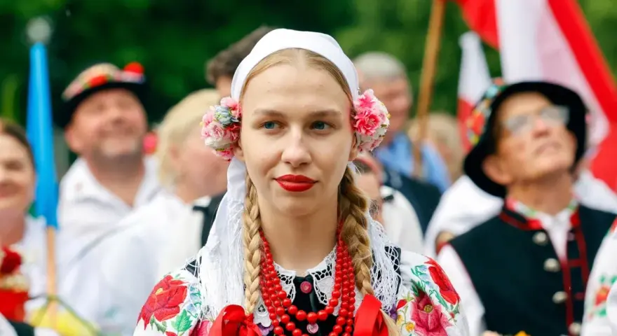 Młoda kobieta w tradycyjnym stroju ludowym z kwiatami we włosach i koralami. Obok ludzie w strojach, biało-czerwone flagi. Świętujemy 16 sierpnia!