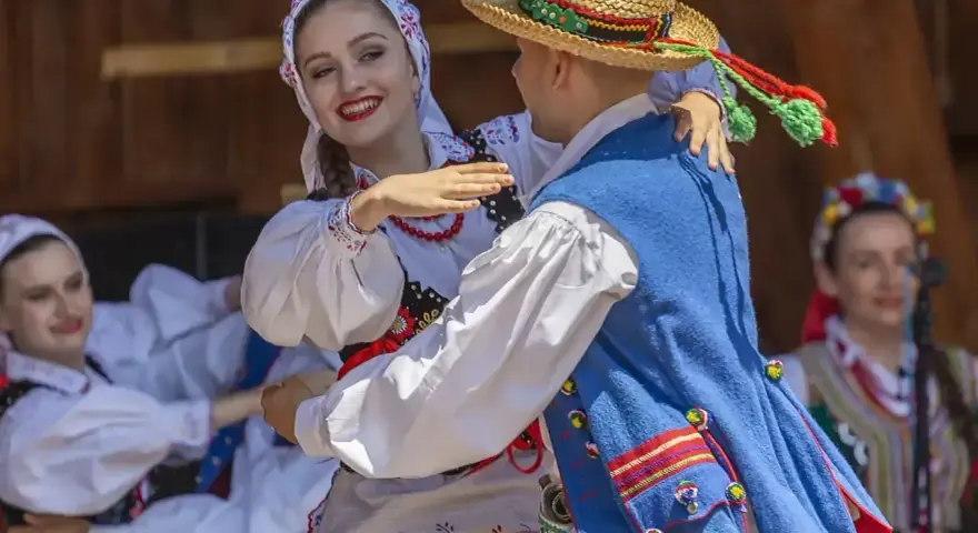 Młoda para w strojach ludowych tańczy z uśmiechem. Chłopak w słomkowym kapeluszu, dziewczyna w kwiecistej chustce. Licho co to za piękny widok!