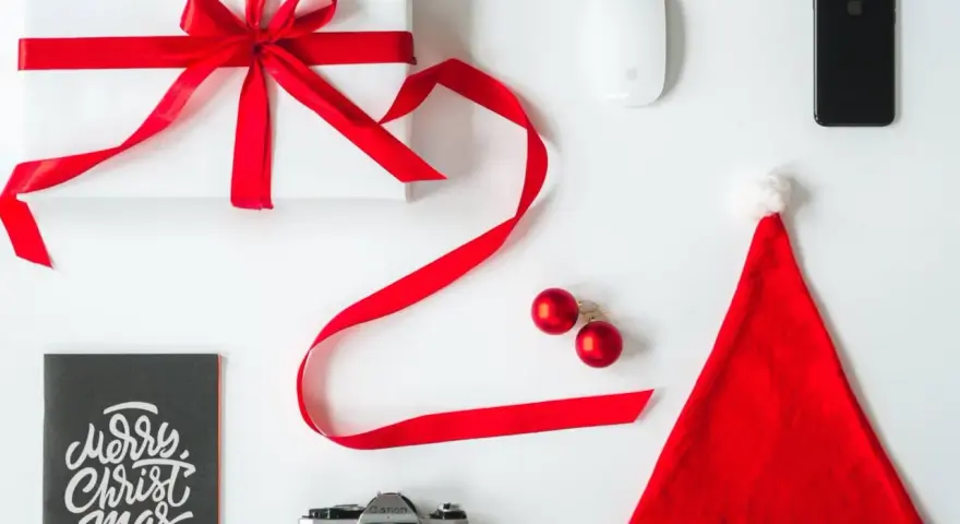 Świąteczne prezenty dla informatyka: białe pudełko z czerwoną wstążką, myszka, iPhone, aparat, kartka "Merry Christmas" i czapka Mikołaja.