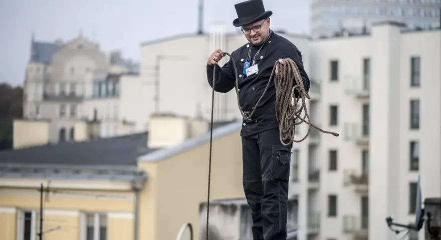 Kominiarz w cylindrze i czarnym stroju przygotowuje się do pracy, trzymając linę. To jego dzień, dzień kominiarza.