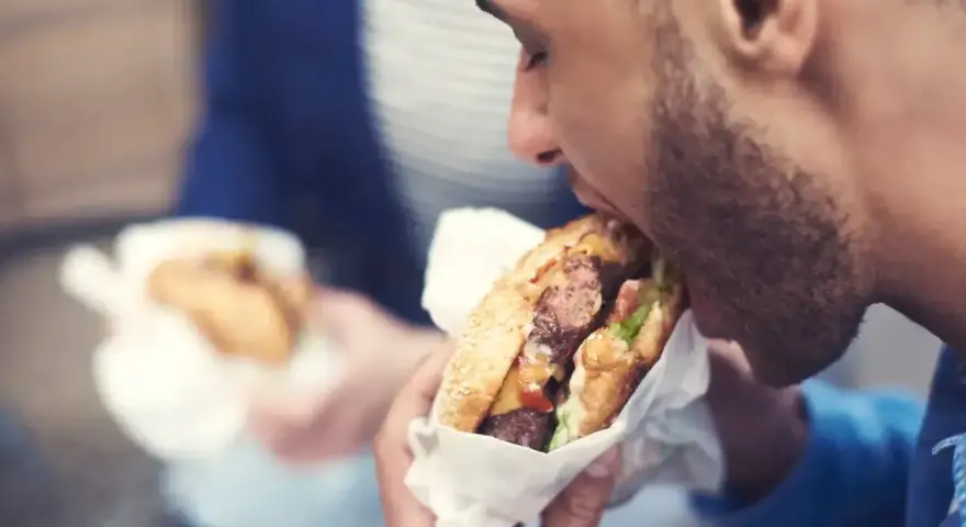 Mężczyzna z zarostem zajada soczystego burgera, świętując dzień burgera. W tle widać drugą osobę z podobnym przysmakiem.