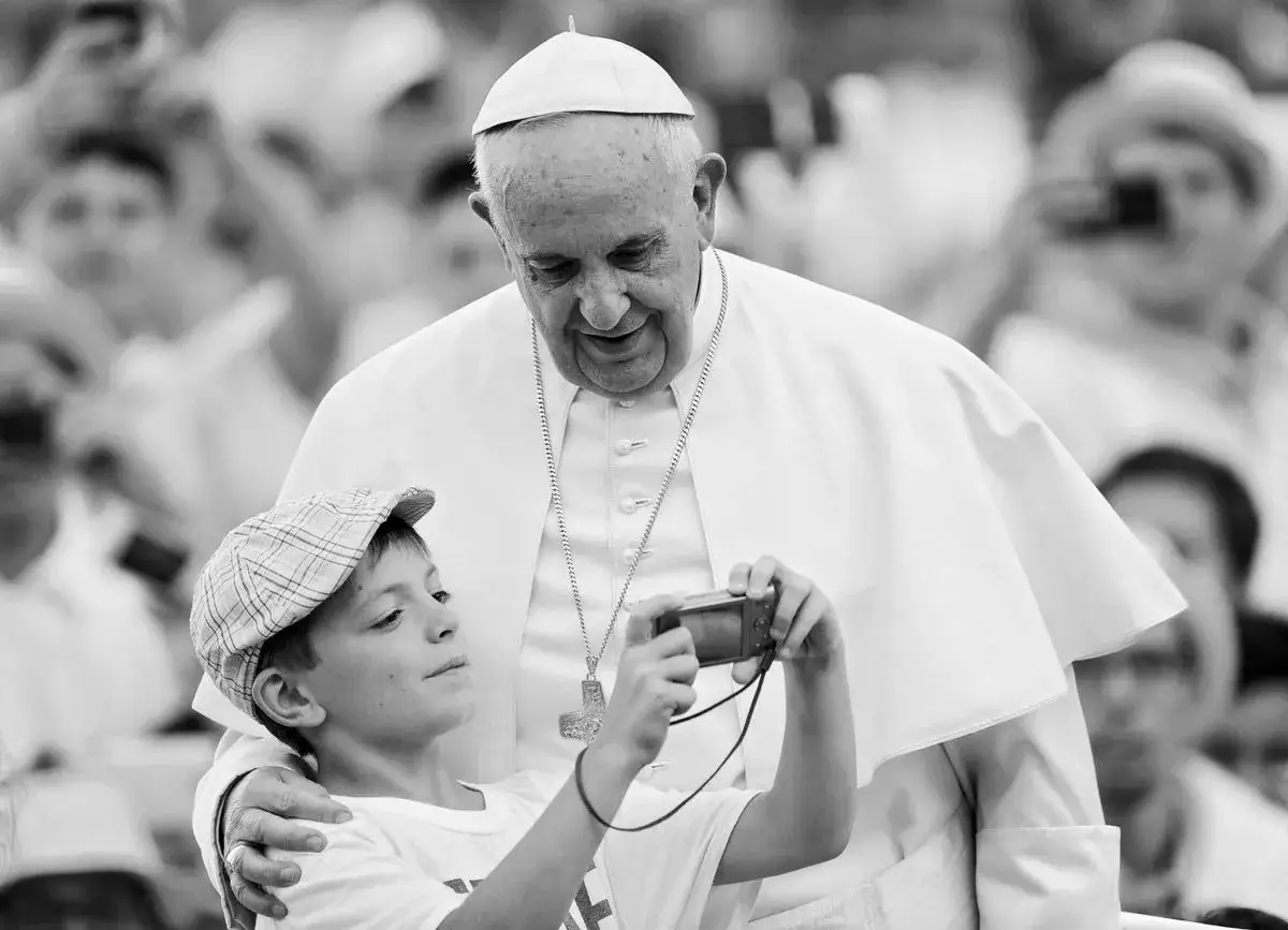 Chłopiec w czapce robi zdjęcie Papieżowi Franciszkowi. W tle tłum ludzi, może to uroczystość z okazji imieniny Franciszka.